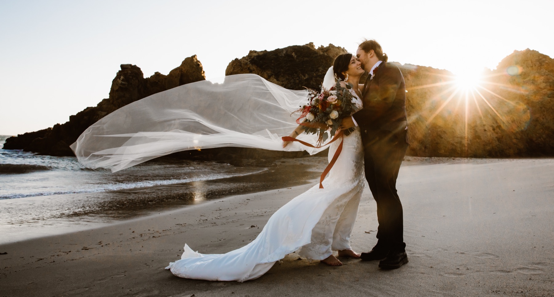 Couple kissing on Big Sur beach with wind-blown veil and sun flare.